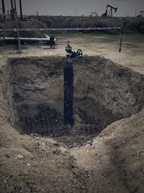 Wellhead casing exposed in excavation pit, danger tape marking the work area